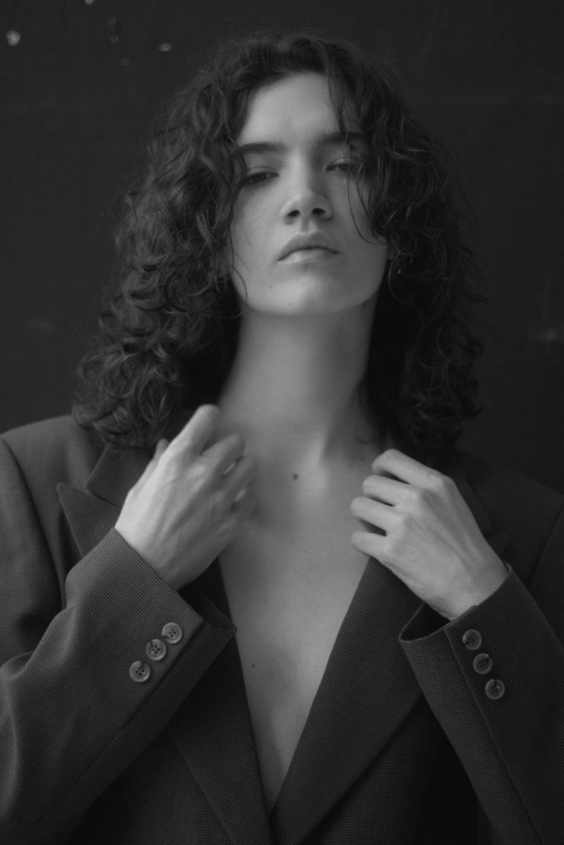 Portrait of a woman with dark curly hair wearing a dark tailored blazer against a dark background.
