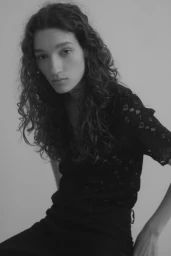 Portrait of a model with long curly hair wearing a dark patterned top against a neutral light grey background.
