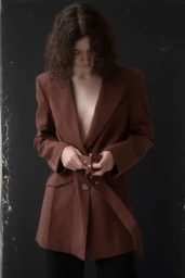 A model with dark curly hair wearing a brown belted jacket against a dark textured background.