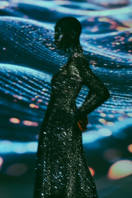 Monochromatic shot of an adult female in a shimmering black gown, posing against a blurred blue and white backdrop with bokeh lights.