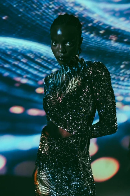 Adult woman in a sparkly high-neck dress with long sleeves, against an abstract blue and white background with bokeh lights.