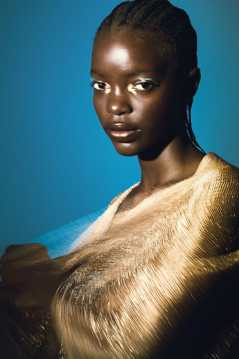 Portrait of a Black woman with braided hair and blue eyeliner, wearing a gold textured garment against a turquoise background.