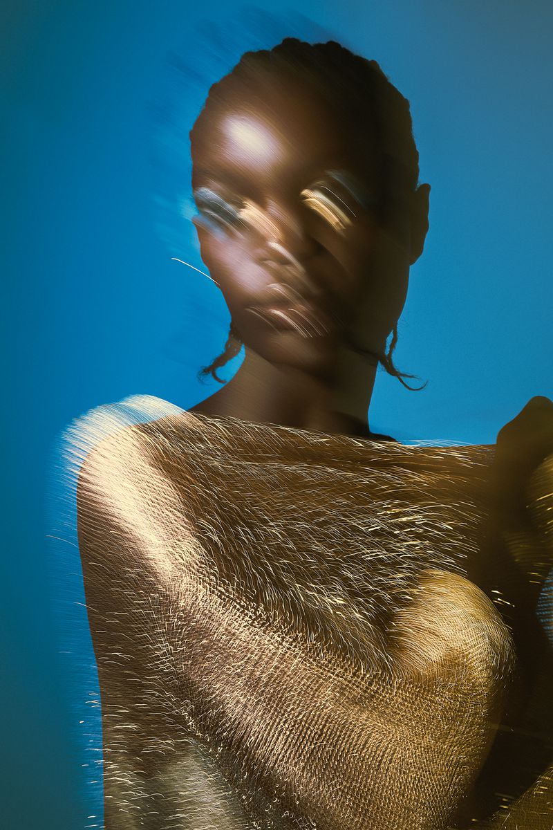 Close-up portrait of a woman with blurred motion effect, adorned with shimmering gold fabric against a bright blue background.