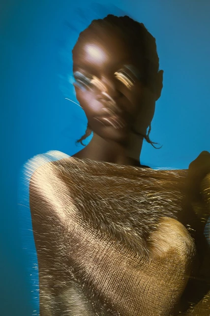 Close-up portrait of a woman with blurred motion effect, adorned with shimmering gold fabric against a bright blue background.