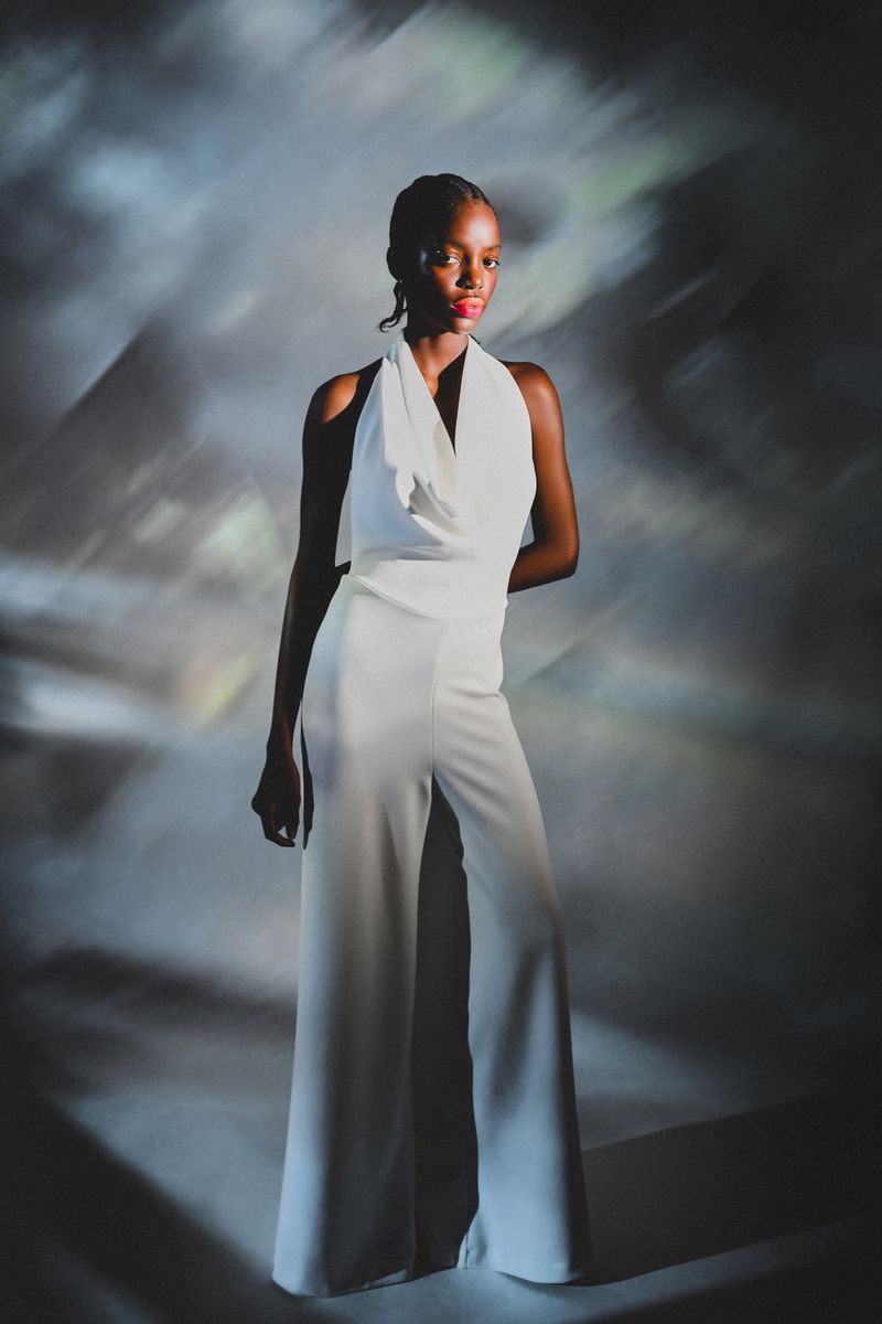 Elegant woman in a white halter jumpsuit posing in a studio setting, with soft, abstract lighting and a neutral background, fashion portrait.