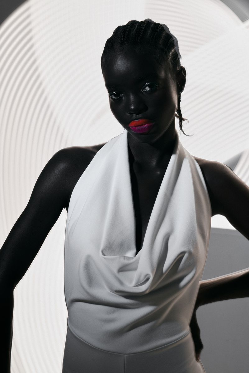 Dark-skinned model in white halter top, posing against an abstract white background with diffused lighting in a studio fashion shot.