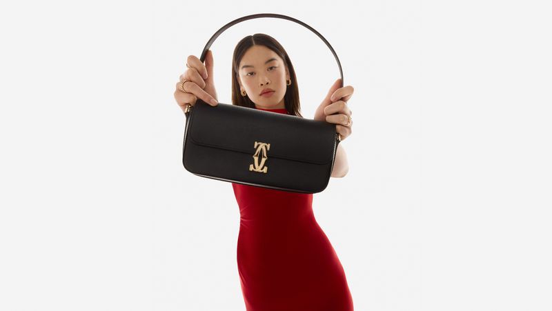 An Asian woman in a red dress poses against a white background, holding a black leather handbag with a gold logo.