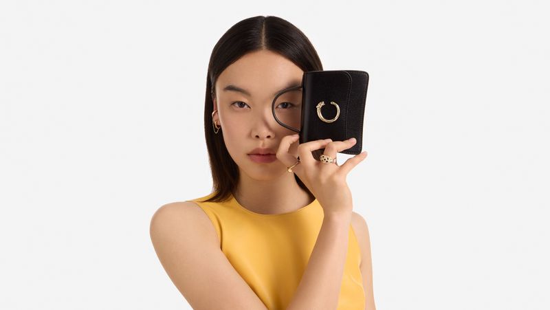 Young woman in yellow top holds black wallet with gold ring detail against a bright white studio backdrop for fashion shot.