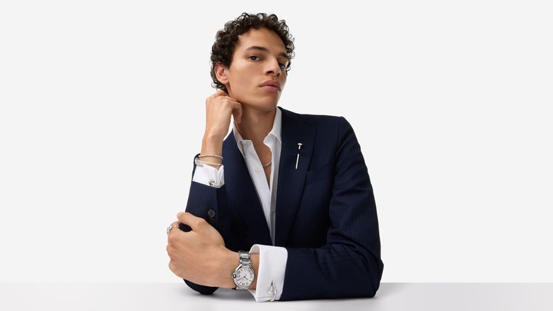 Man in suit poses indoors. He is wearing a watch, bracelet, and ring. The setting is minimalist with a white background, emphasizing the subject.