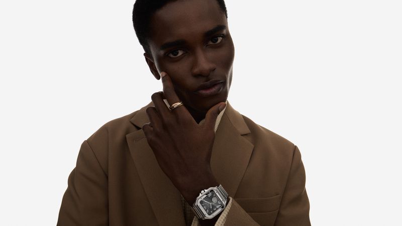 Close-up of a dark-skinned male model in a tan blazer, hand on face, showcasing a wristwatch with a silver band against a white backdrop.
