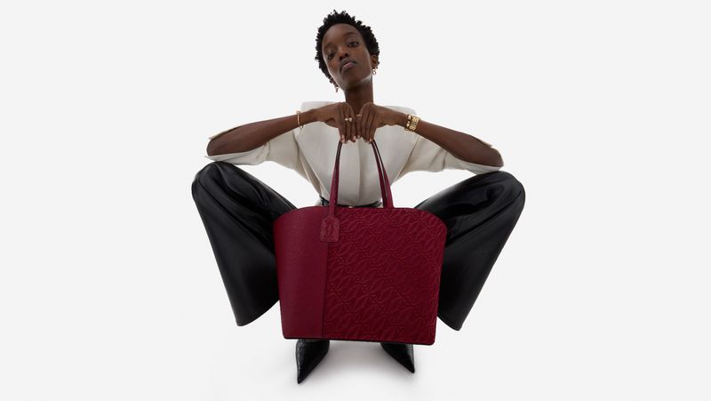Dark-skinned woman poses on a white backdrop, seated with legs spread, holding a dark red patterned leather tote bag.