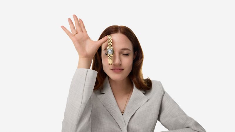Elegant adult woman in grey business jacket posing with a luxury gold watch over her eye in a bright indoor studio setting.