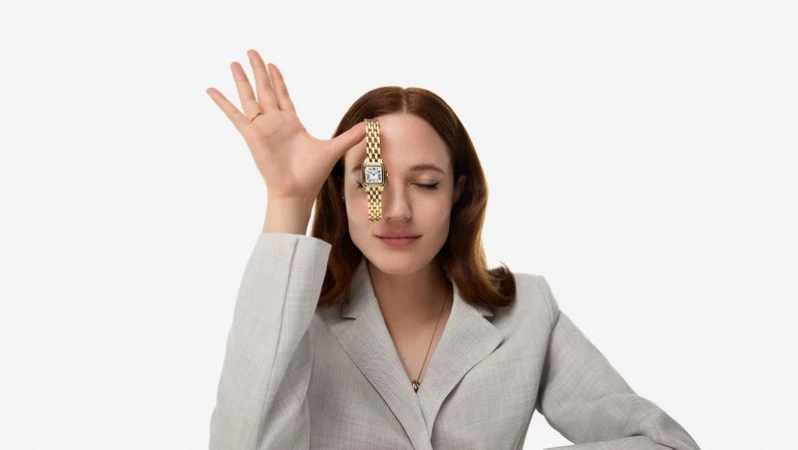 Elegant adult woman in grey business jacket posing with a luxury gold watch over her eye in a bright indoor studio setting.