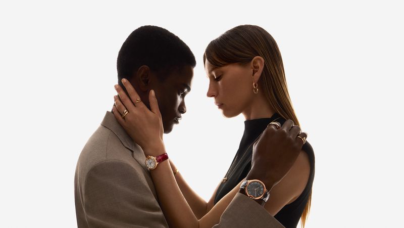 A stylish interracial couple embraces in a bright studio, showcasing wristwatches and gold jewelry against a clean white backdrop.