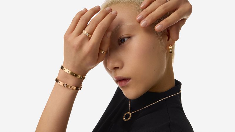 Portrait of a woman with short blonde hair wearing gold jewelry against a white background, including rings, bracelets, necklace, and earrings.
