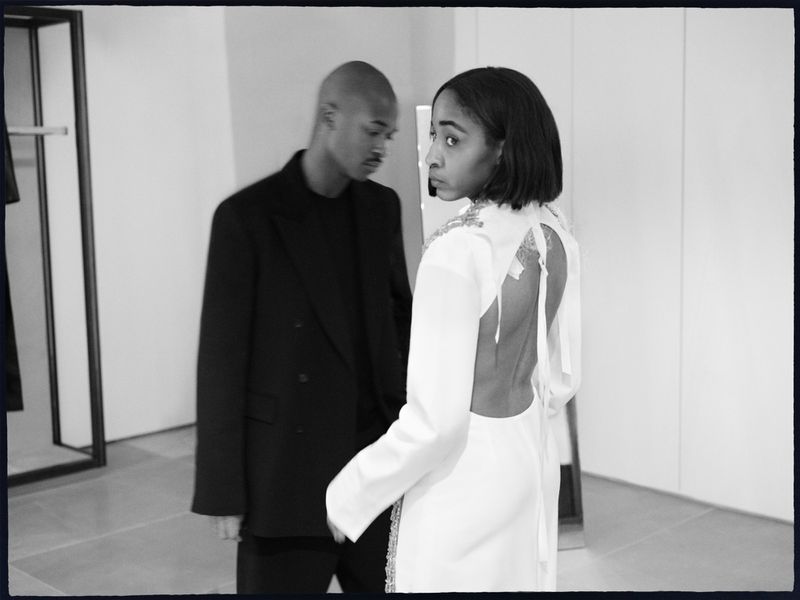 A woman in a white dress with an open back looks over her shoulder while a man in a black suit stands in the background indoors.