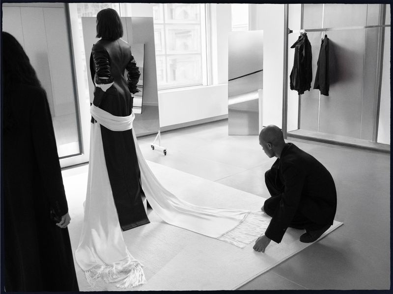 Black and white editorial image showing a person standing in a gown with a long white train being adjusted by someone kneeling.