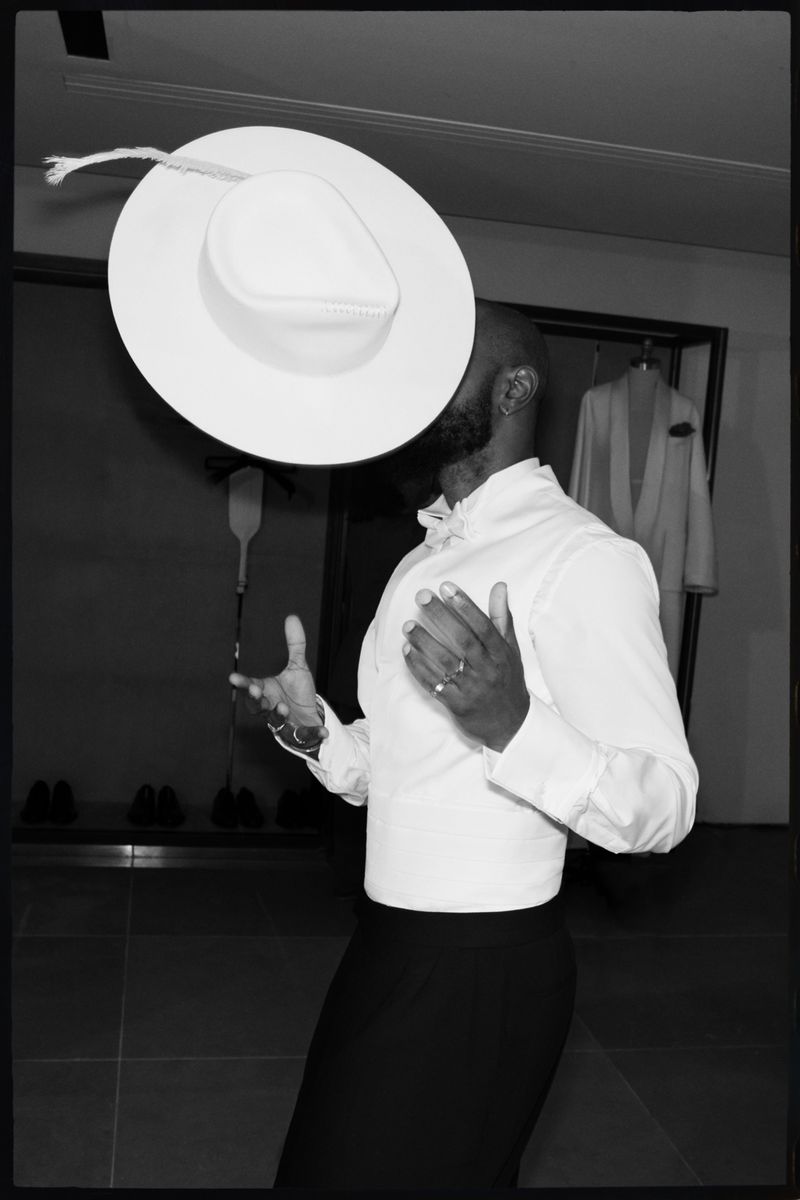 Black and white portrait of a Black adult male in a collared shirt, with a large white hat hovering above his head in an indoor space.