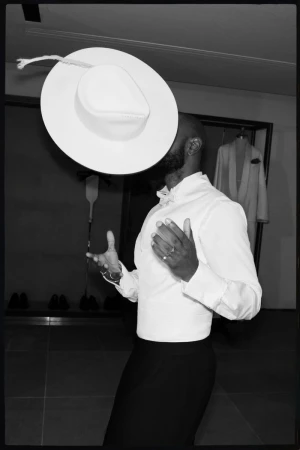 Black and white portrait of a Black adult male in a collared shirt, with a large white hat hovering above his head in an indoor space.