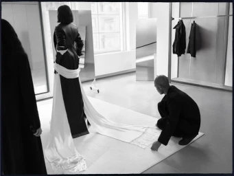 Black and white editorial image showing a person standing in a gown with a long white train being adjusted by someone kneeling.
