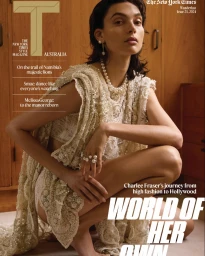 A woman crouching in a textured cream lace dress and pearl jewellery against a wooden cabinet background for T Australia.