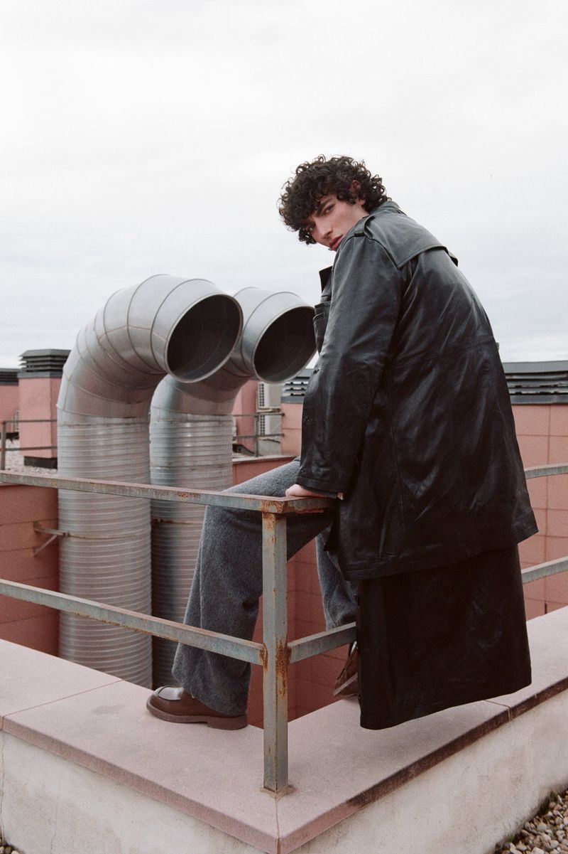 Adult male model in long black coat and wide-legged jeans poses on rooftop with industrial pipes, cool tones, and editorial fashion.