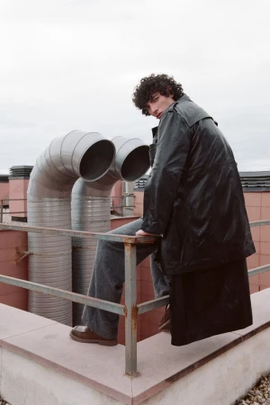 Adult male model in long black coat and wide-legged jeans poses on rooftop with industrial pipes, cool tones, and editorial fashion.