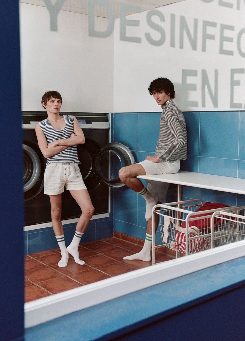Two men in striped tops and shorts pose in a vintage-style laundromat with blue tiled walls and retro washing machines, captured indoors.