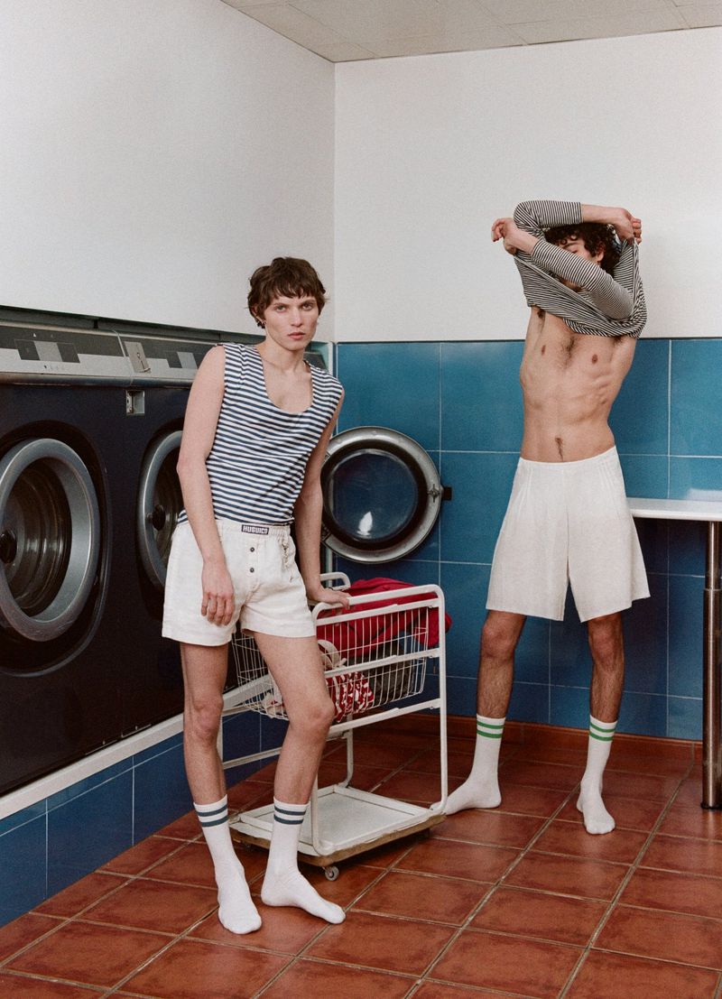 Two adult men in a laundry room. One is wearing shorts and a striped tank top, leaning against a laundry cart. The other is shirtless.