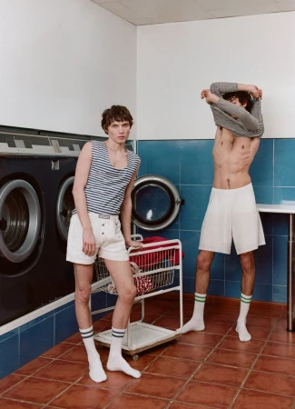 Two adult men in a laundry room. One is wearing shorts and a striped tank top, leaning against a laundry cart. The other is shirtless.