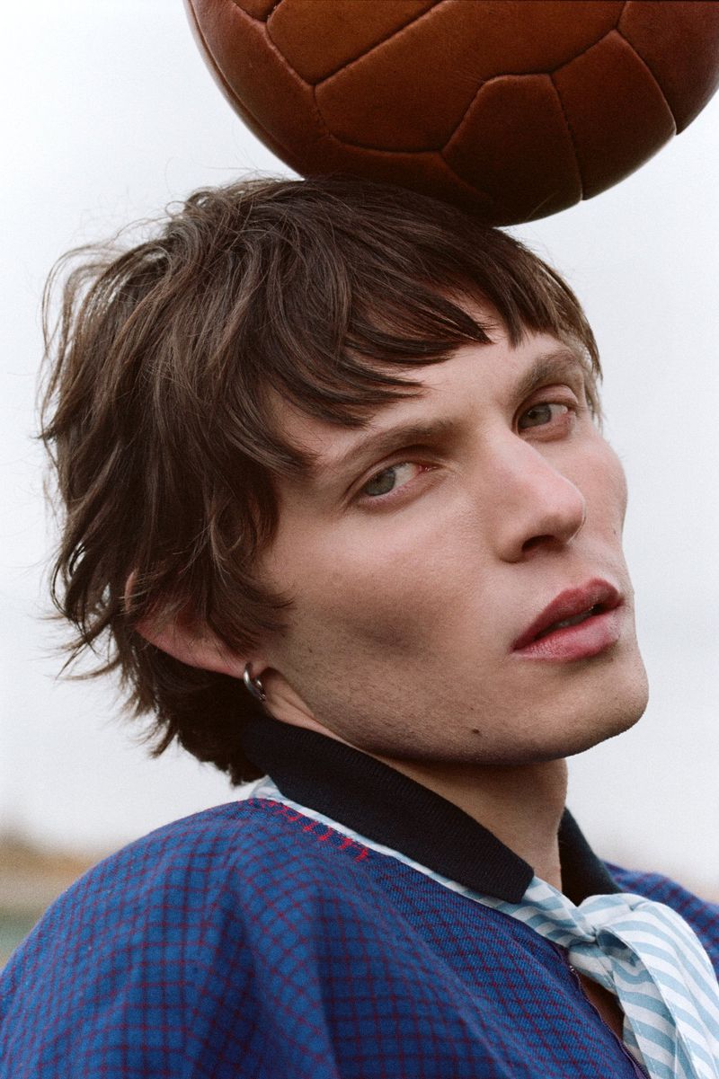 Portrait of a young adult man with dark hair balancing a soccer ball on his head in a blue patterned jacket with a tie.
