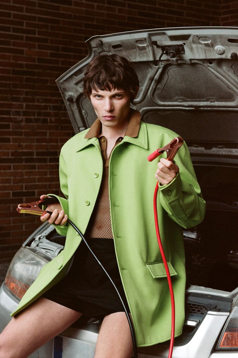 A fashionable young adult man wearing a green coat, sitting on a silver car's hood while holding jumper cables in an urban setting.