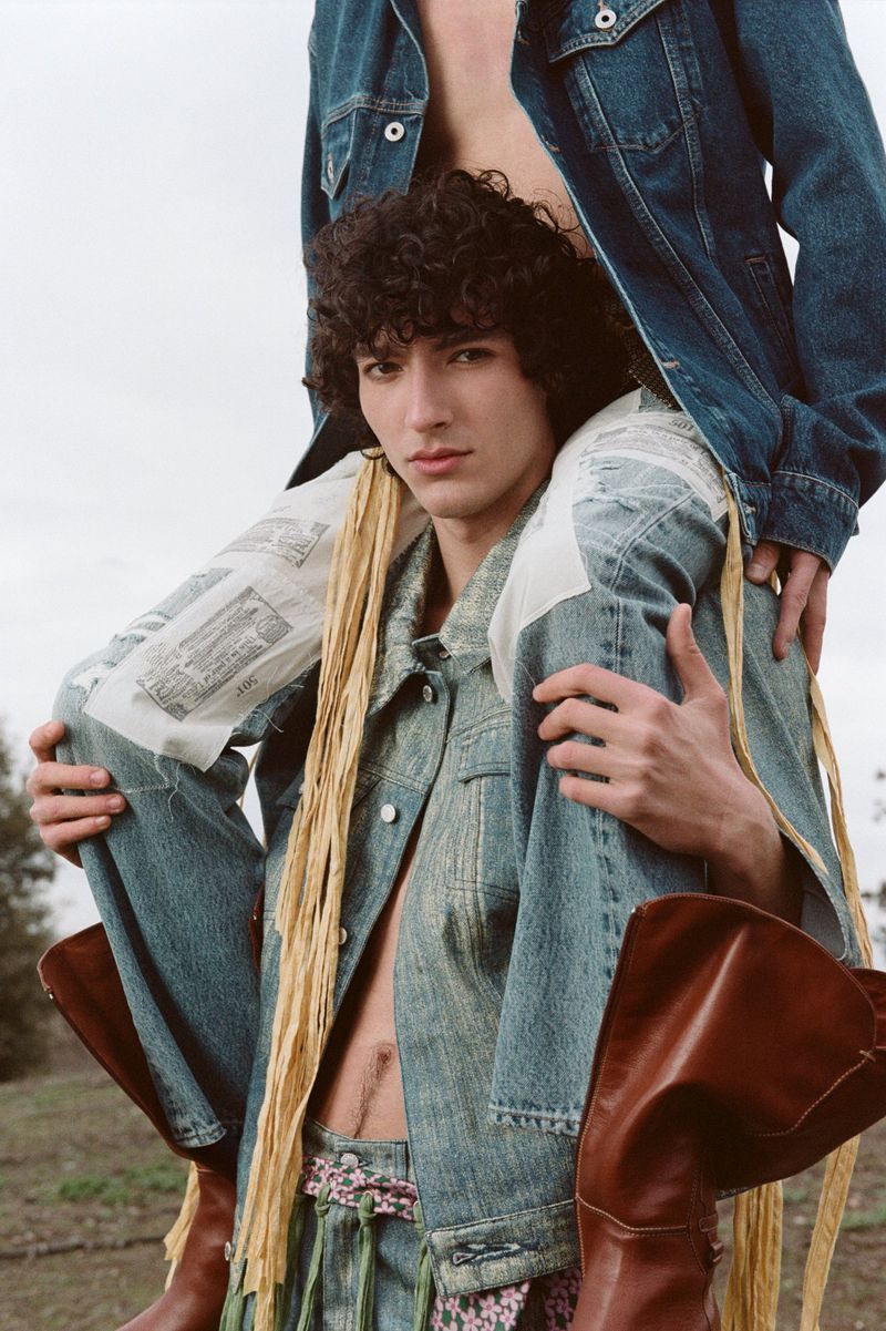 Two men posing outdoors wearing matching denim jackets and pants with fringed details and cowboy boots against a muted background.