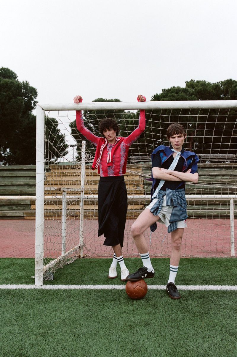 Two young adult males are posing on a soccer field in sports-inspired fashion. The men are standing near a soccer goal with a soccer ball.
