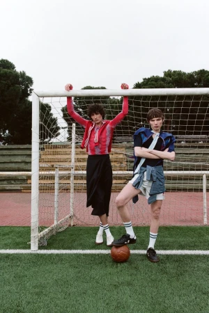 Two young adult males are posing on a soccer field in sports-inspired fashion. The men are standing near a soccer goal with a soccer ball.