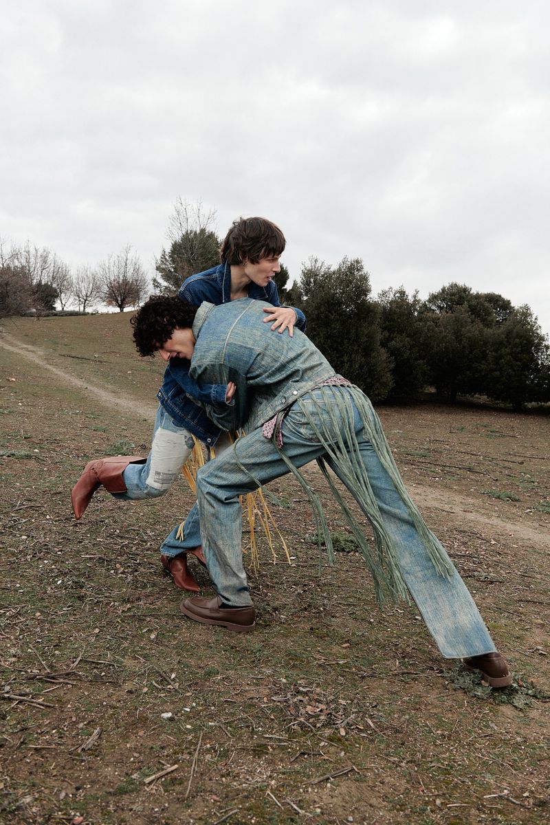 Two people in denim outfits playfully wrestle on a grassy hillside with sparse trees under an overcast sky, captured in a medium shot.
