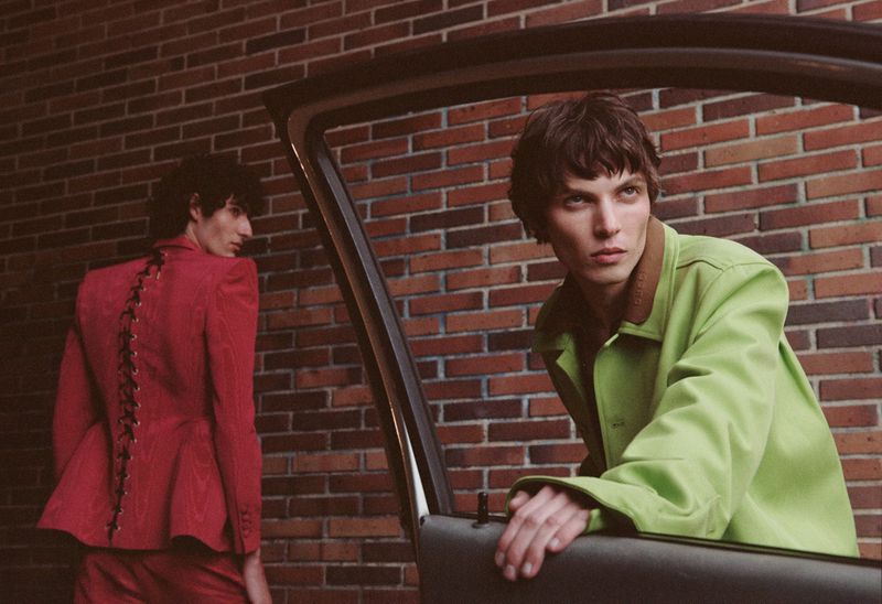 Two dark-haired young men in fashion blazers pose in front of a brick wall next to a car; the primary model looks to the side.