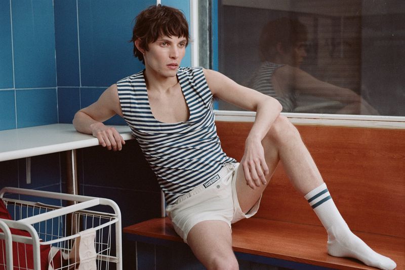 Male fashion model poses indoors wearing a striped tank top, white shorts, and striped socks while sitting on a wooden bench.