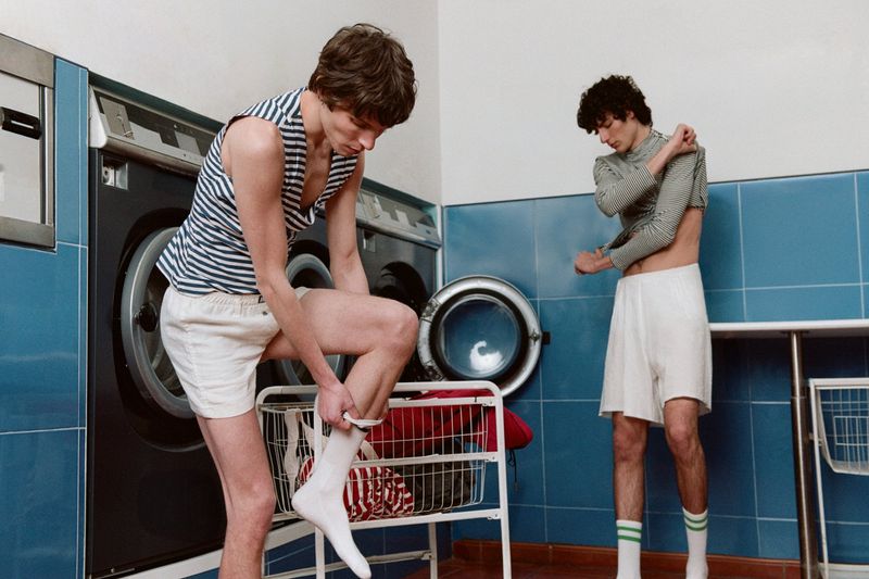 Two young adult men in a laundry room. One man wears a striped tank and shorts, pulling on socks; the other adjusts his shirt.