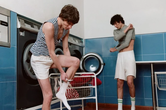 Two young adult men in a laundry room. One man wears a striped tank and shorts, pulling on socks; the other adjusts his shirt.