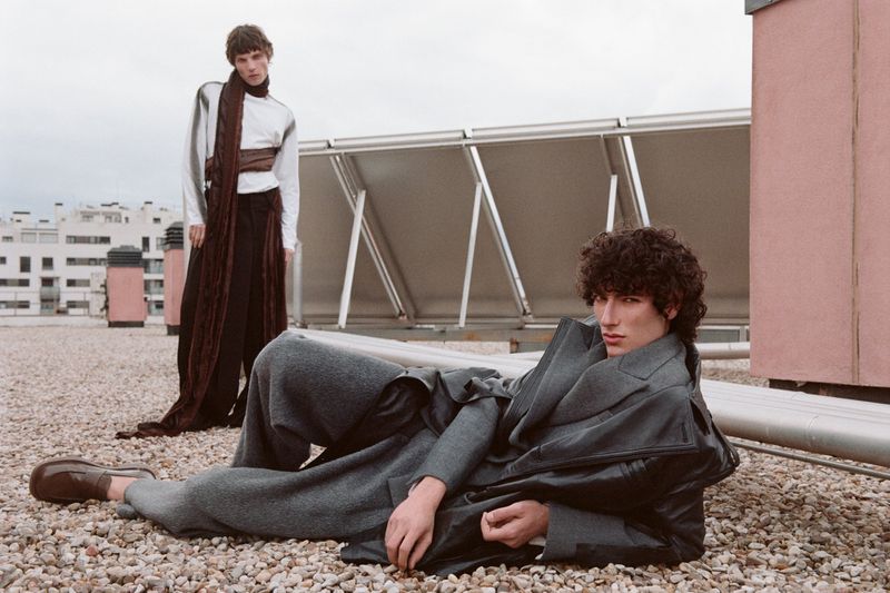 Two adult males model fashion on a rooftop with solar panels in the background, one lying down and one standing; overcast daylight.