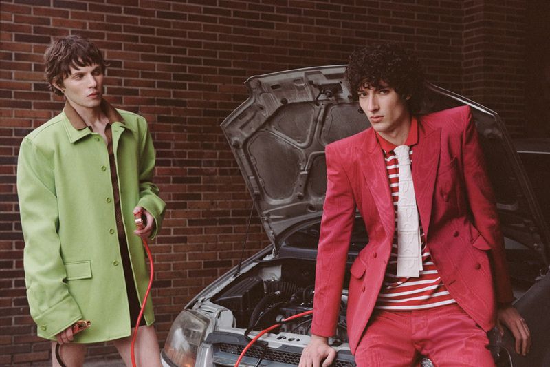 Two adult males in brightly colored suits pose next to a car with its hood open against a brick wall background outdoors.
