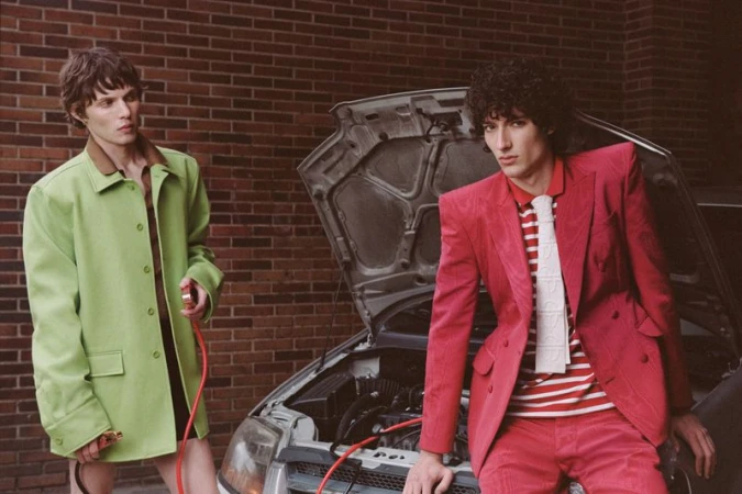 Two adult males in brightly colored suits pose next to a car with its hood open against a brick wall background outdoors.