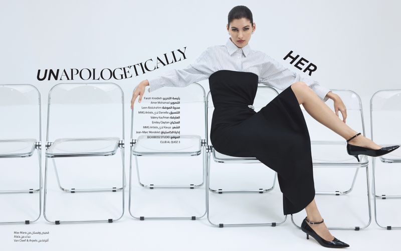 A woman in a black dress and white shirt is seated on clear chairs in a studio, showcasing a minimalistic fashion editorial look.