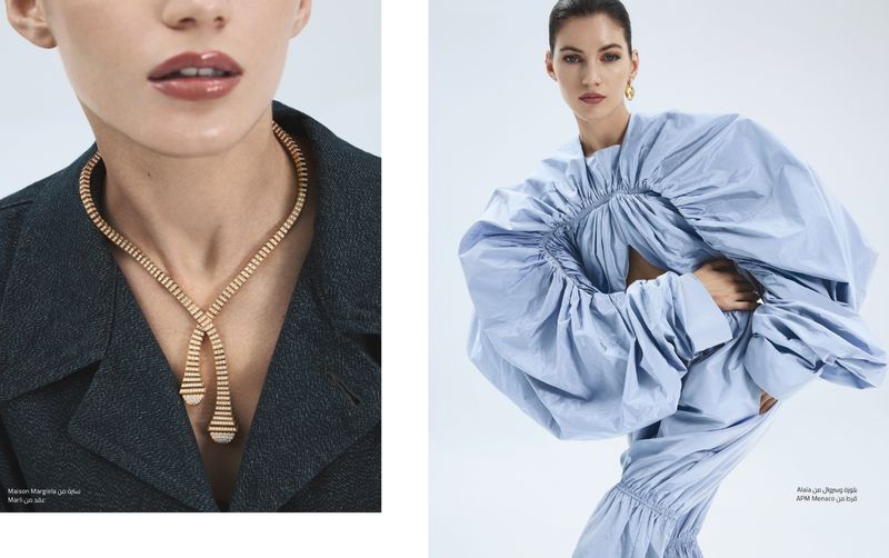Two women showcase light blue and charcoal grey suit jackets. One wears a gold necklace. Minimalist fashion photography with a bright background.