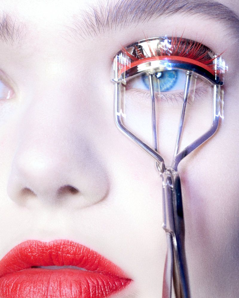 Close-up of a woman's face with bright red lipstick and a metal eyelash curler near one eye, focusing on beauty and makeup.
