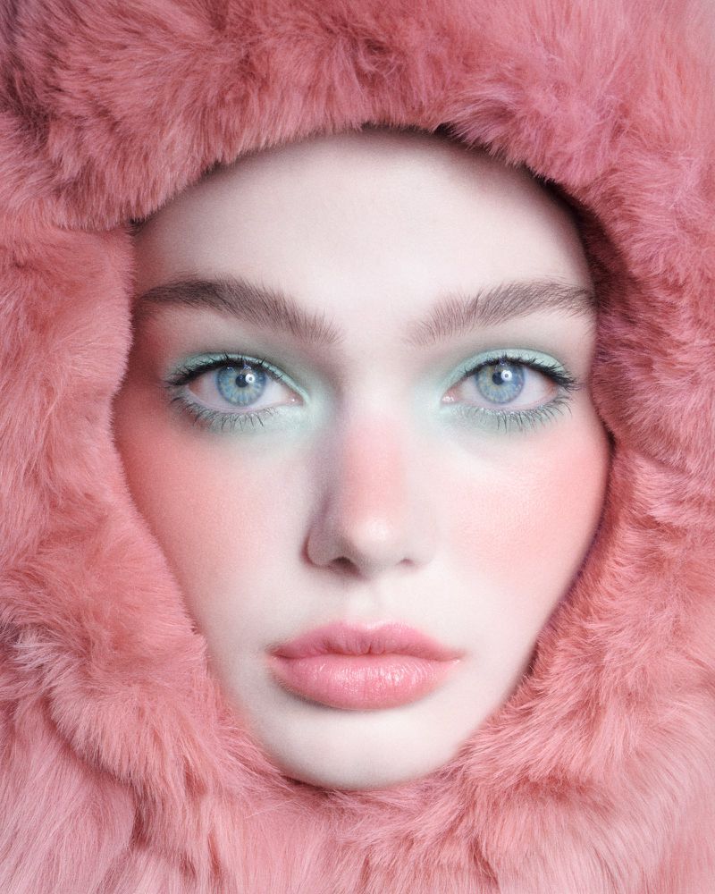 Close-up portrait of a woman with light blue eyes and pink makeup wearing a pink fur hood in a soft, diffused light.