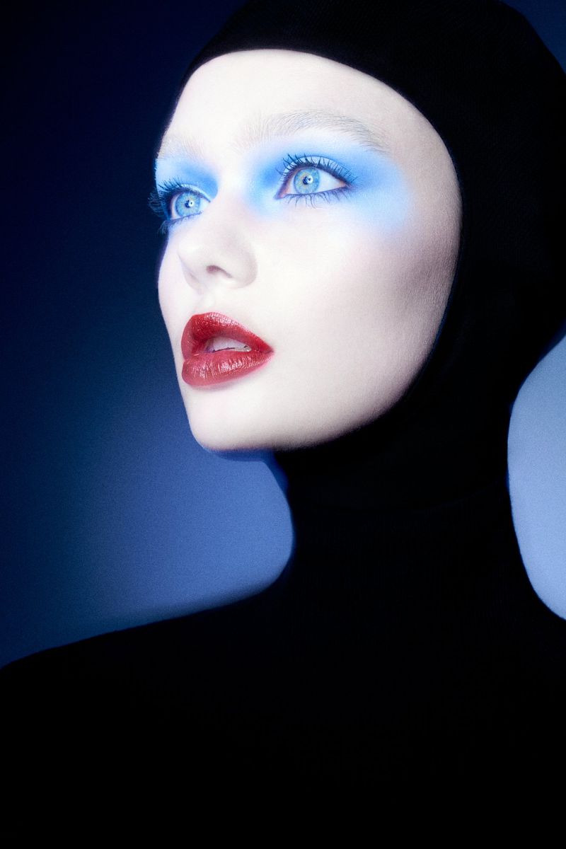 Fashion portrait of an adult female model wearing a black hood, with pale skin, red lipstick, and blue eye makeup on a blue background.