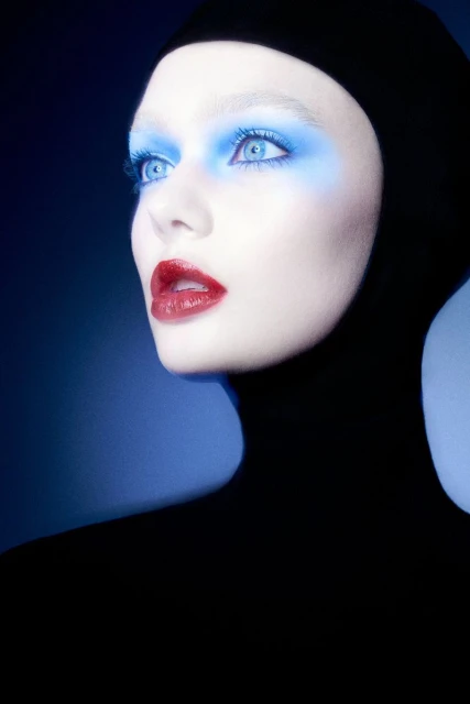 Fashion portrait of an adult female model wearing a black hood, with pale skin, red lipstick, and blue eye makeup on a blue background.