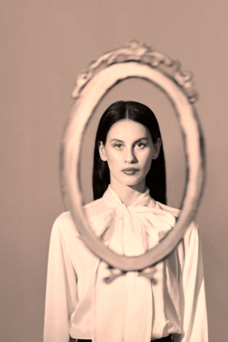 Indoor portrait of a young adult woman looking through an antique mirror in a studio, posing in a vintage fashion style.
